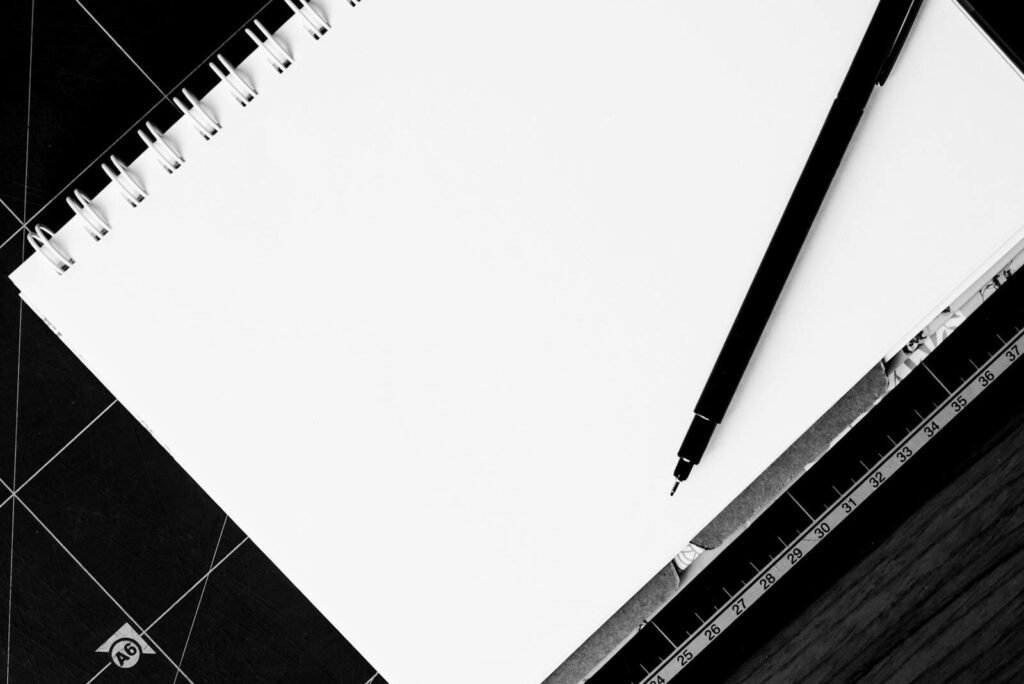 Black and white image of a blank spiral notebook and pen on a ruler-covered surface.